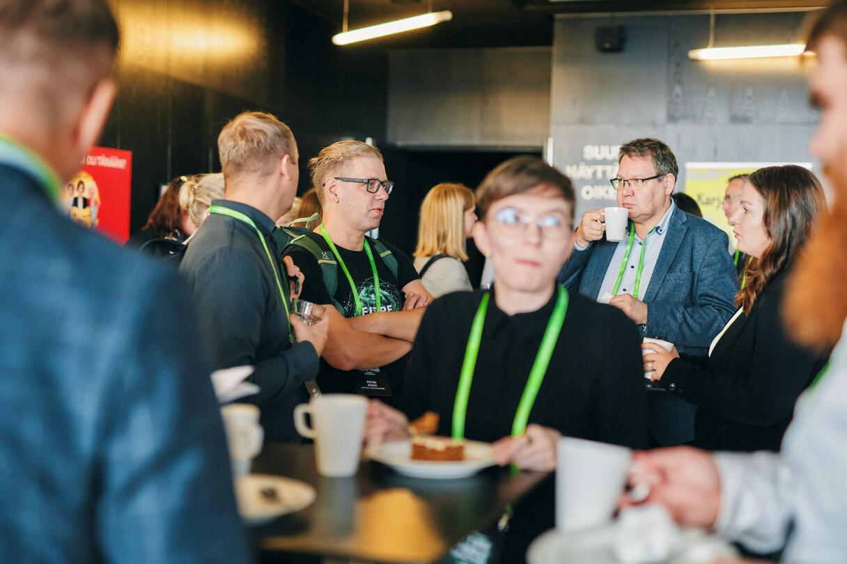People standing and chatting in a lobby-like space while enjoying coffee refreshments, all wearing green lanyards.