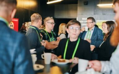 People standing and chatting in a lobby-like space while enjoying coffee refreshments, all wearing green lanyards.